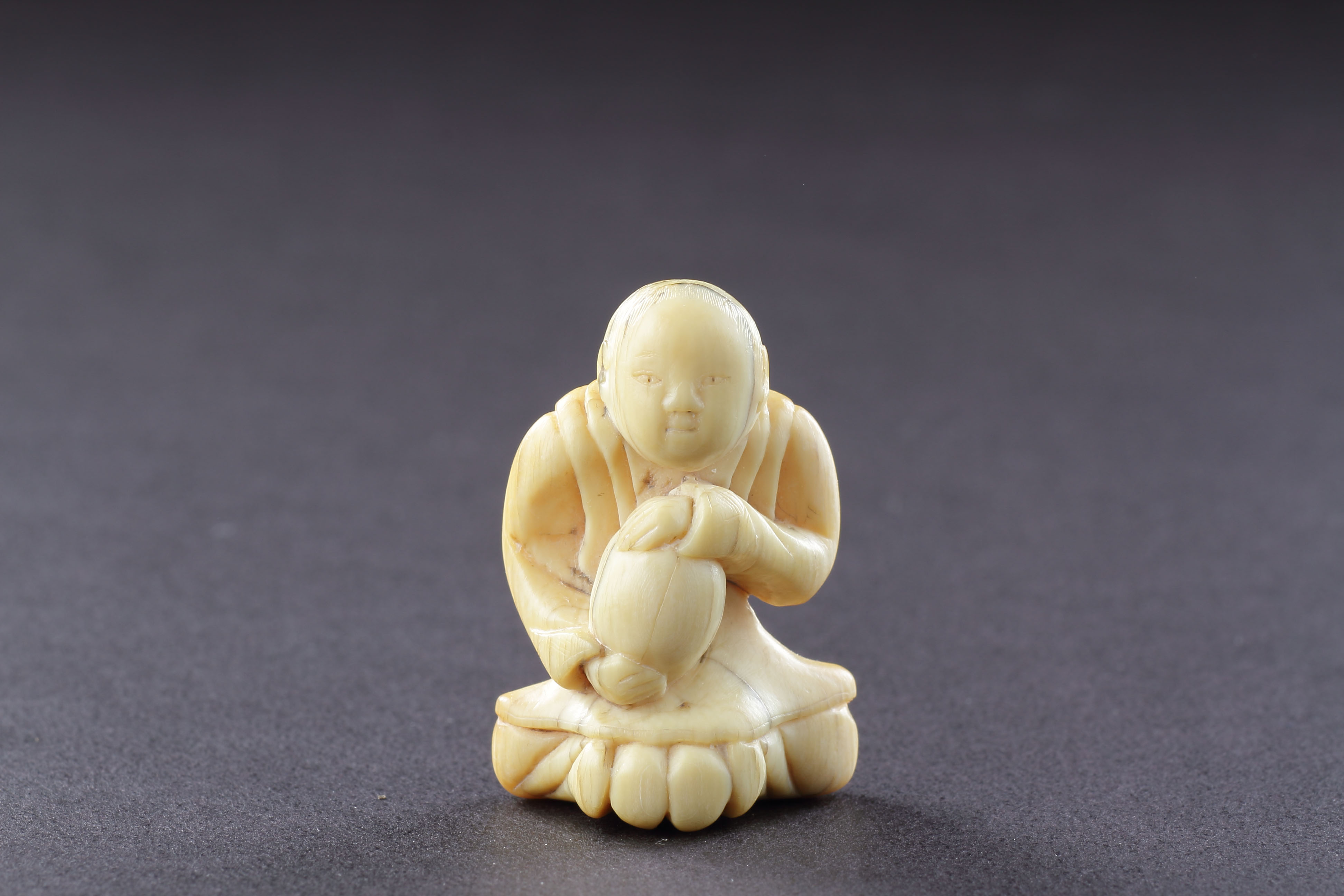 Japanese Ivory Netsuke of a Man Seated Wearing a Mon Decorated Kimono Clasping with Both Hands his Treasured Saké Gourd